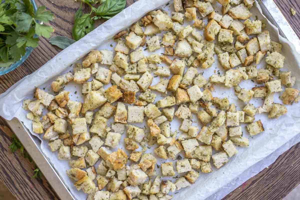 Gluten free croutons on a parchment lined baking sheet with fresh herbs around the pan.