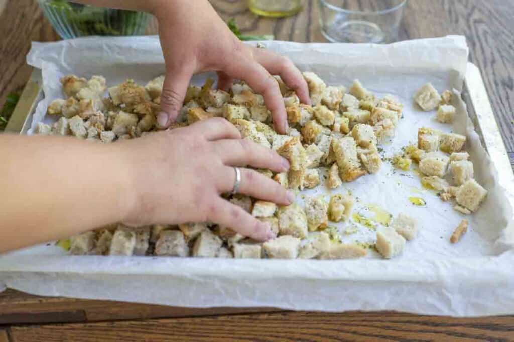 hands combining diced bread with olive on and spices on a parchment lined baking sheet