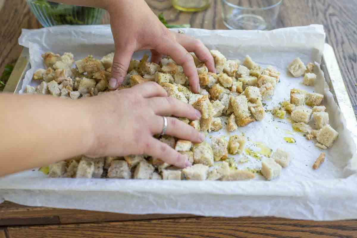 Combining diced bread with olive oil and spices on a parchment lined baking sheet.
