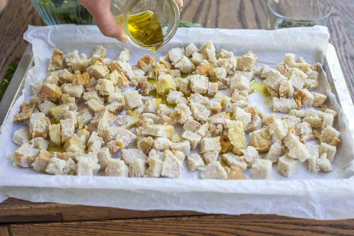 Pouring olive oil over diced bread on a parchment lined baking sheet.