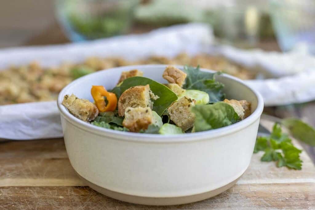 cream colored bowl filled with salad and topped with homemade gluten free croutons on a wooden table. More croutons on a parchment lined baking sheet are in the background