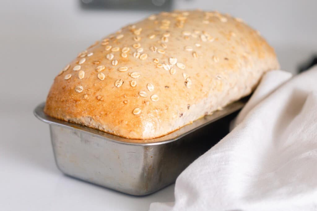 loaf of multigrain sourdough bread in a loaf pan on a white countertop with a white towel to the right