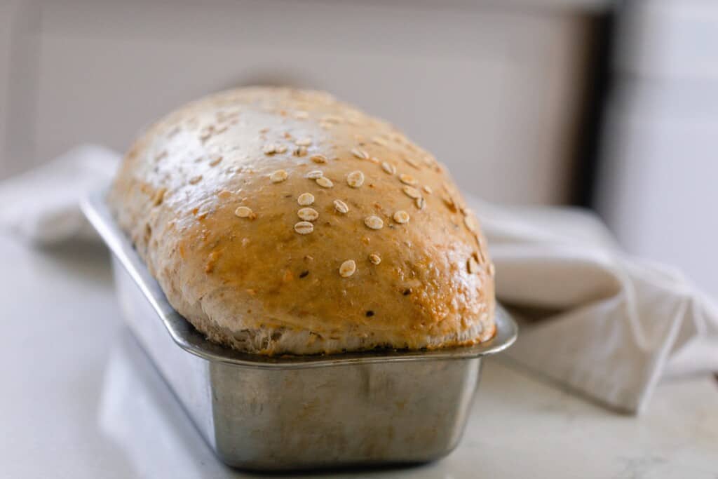 loaf of soft multigrain sourdough bread topped with oats in a loaf pan on a white countertop