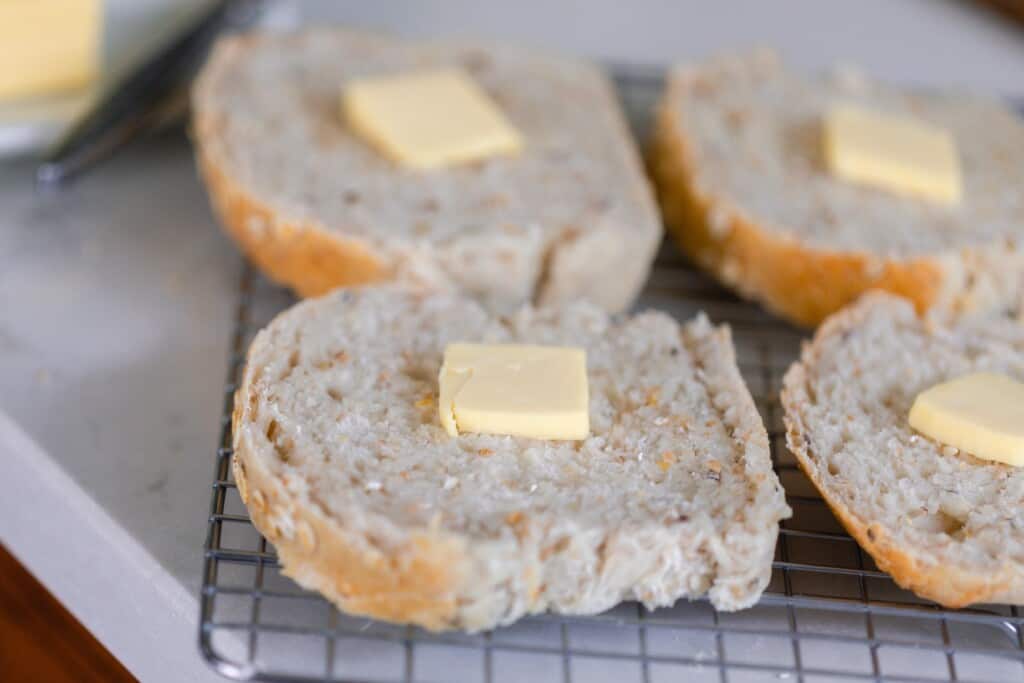 four slices of multigrain sourdough with a tablespoon of butter on each all sitting on a black cooling rack on a white kitchen counter