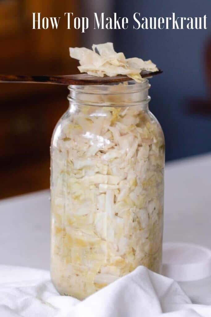 half gallon mason jar full of homemade sauerkraut with a wooden spoon with a serving of sauerkraut resting on the top of the jar. The jar its on a white countertop with a white towel surrounding the jar