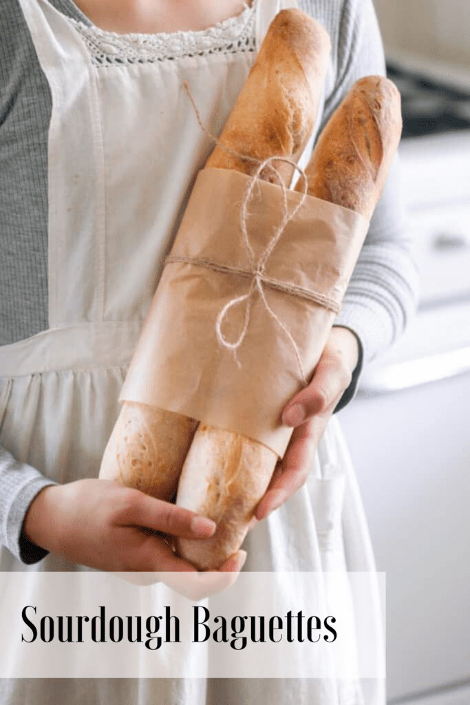 Girl in a white apron holding two loaves of sourdough baguettes wrapped in brown parchment paper and tied with a string