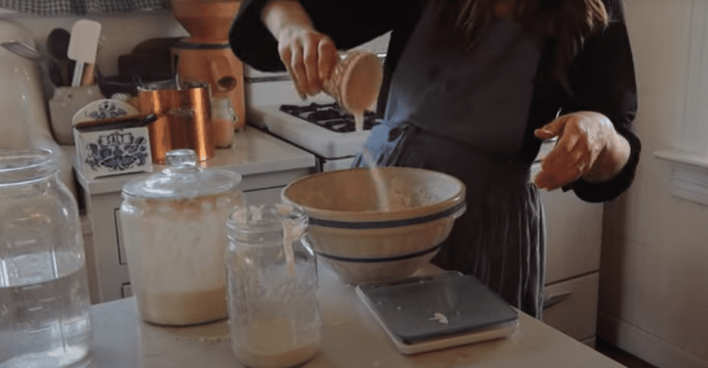 Woman in blue apron adding salt to dough in mixing bowl with kitchen scale nearby