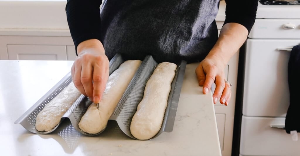 someone scoring three loaves of sourdough baguettes before putting them in an oven