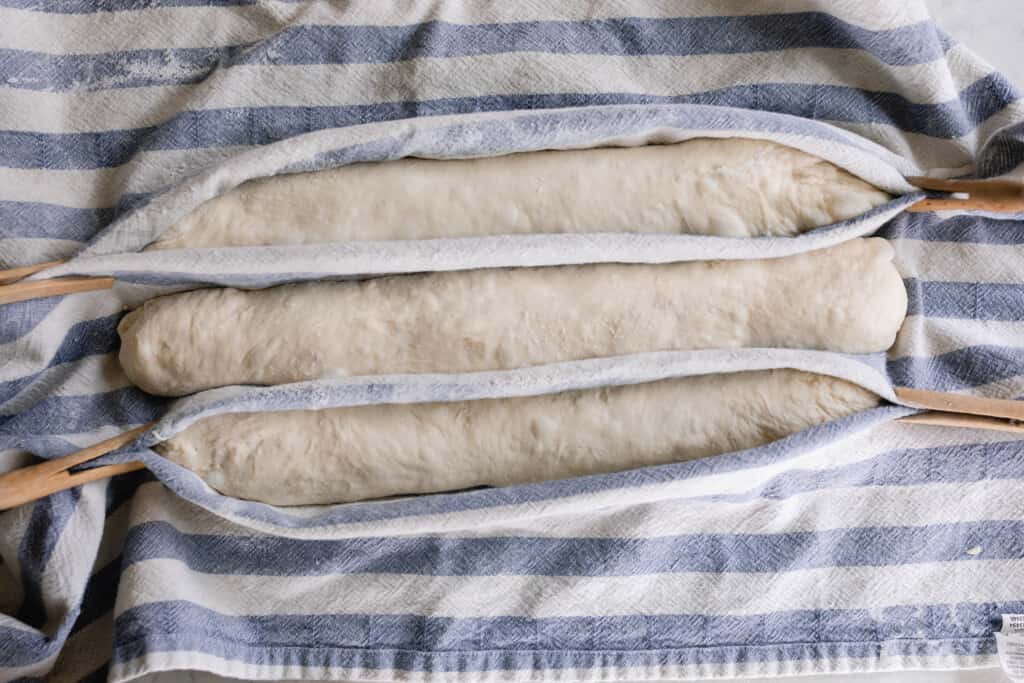 sourdough baguettes shaped and in a baker's couche made of a white and blue striped tea towel