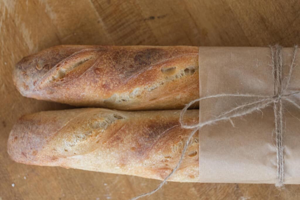 two loaves of sourdough baguettes wrapped in parchment and twine on a wood cutting board