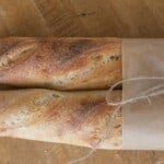 two loaves of sourdough baguettes wrapped in parchment and twine on a wood cutting board