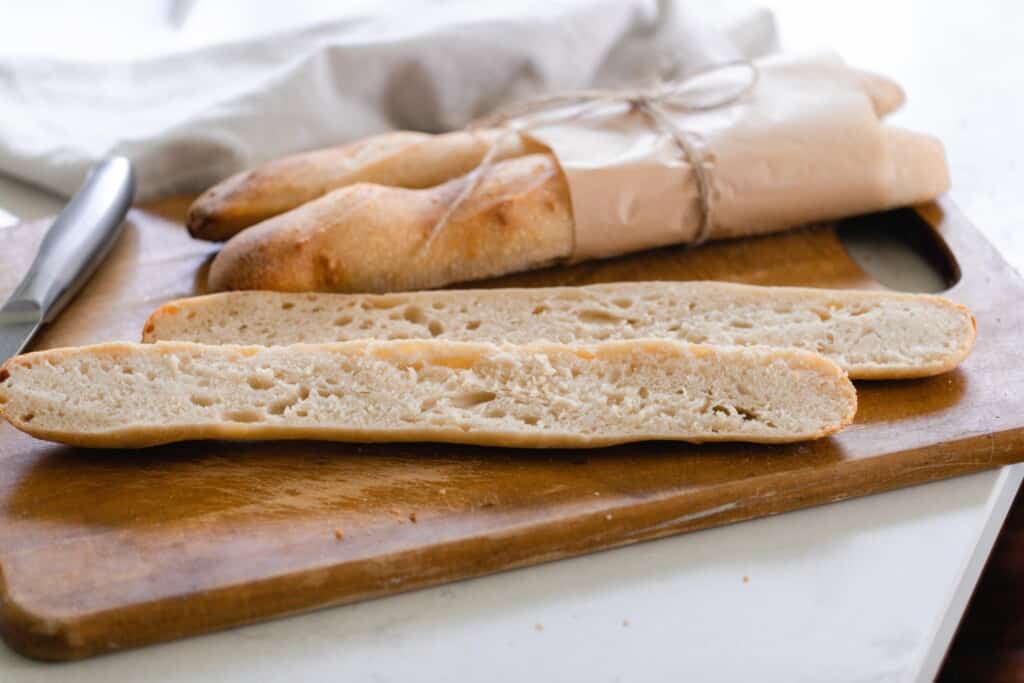 Two sourdough baguettes in brown parchment paper with one baguette sliced open on a cutting board