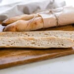 Two sourdough baguettes in brown parchment paper with one baguette sliced open on a cutting board