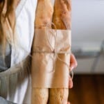 girl wearing a white apron holding two sourdough baguettes wrapped in parchment paper and twine.