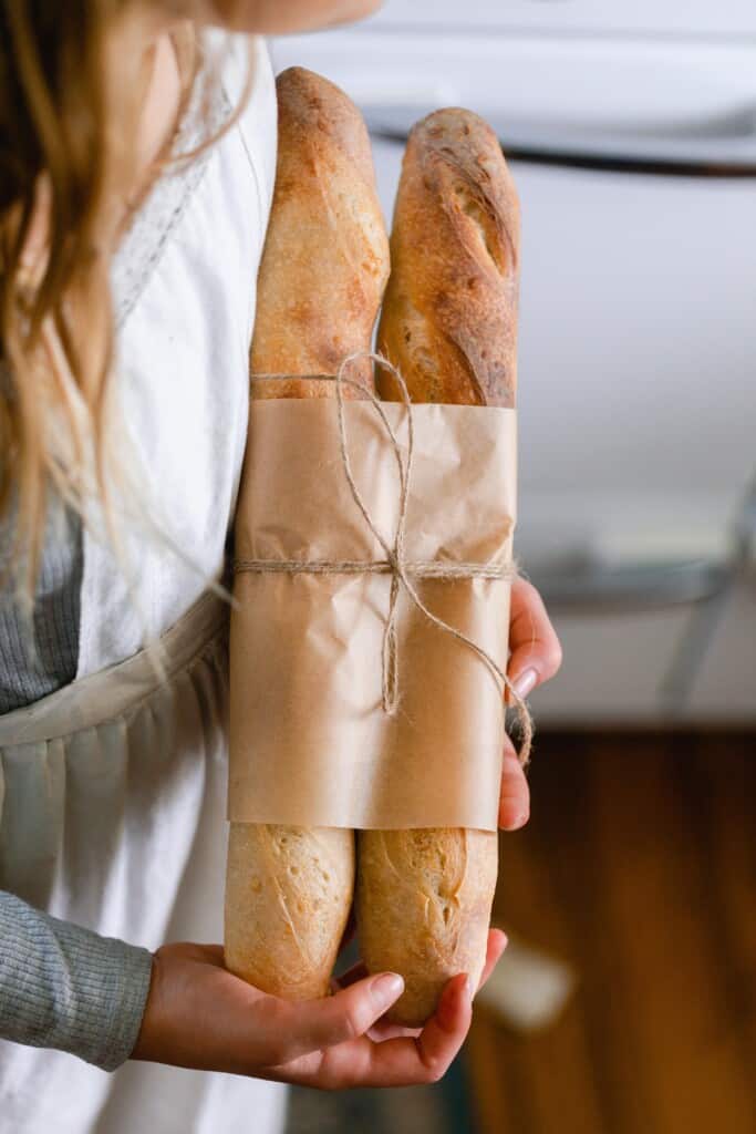 girl wearing a white apron holding two sourdough baguettes wrapped in parchment paper and twine.