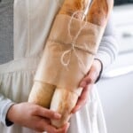 someone holding two baguettes wrapped in brown parchment paper.