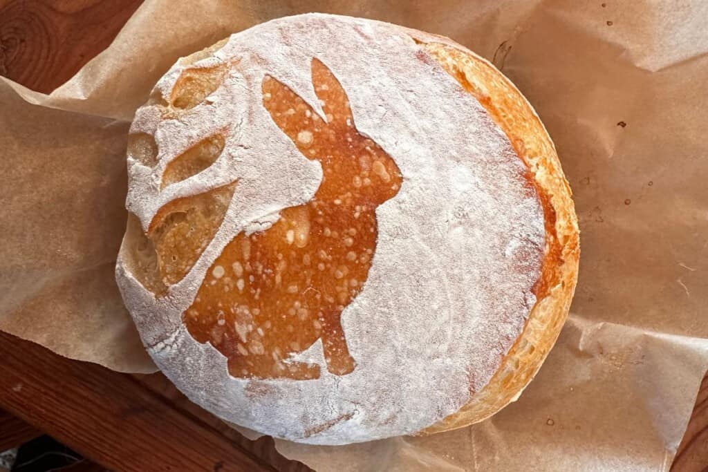 boule of sourdough bread with a bunny and wheat design on top of the loaf. The loaf is on parchment paper