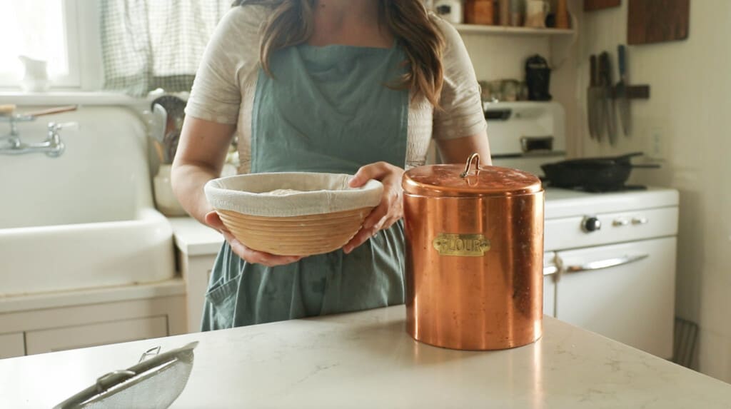 a woman wearing a green apron holding banneton basket with a copper canister in front of her own a white countertop