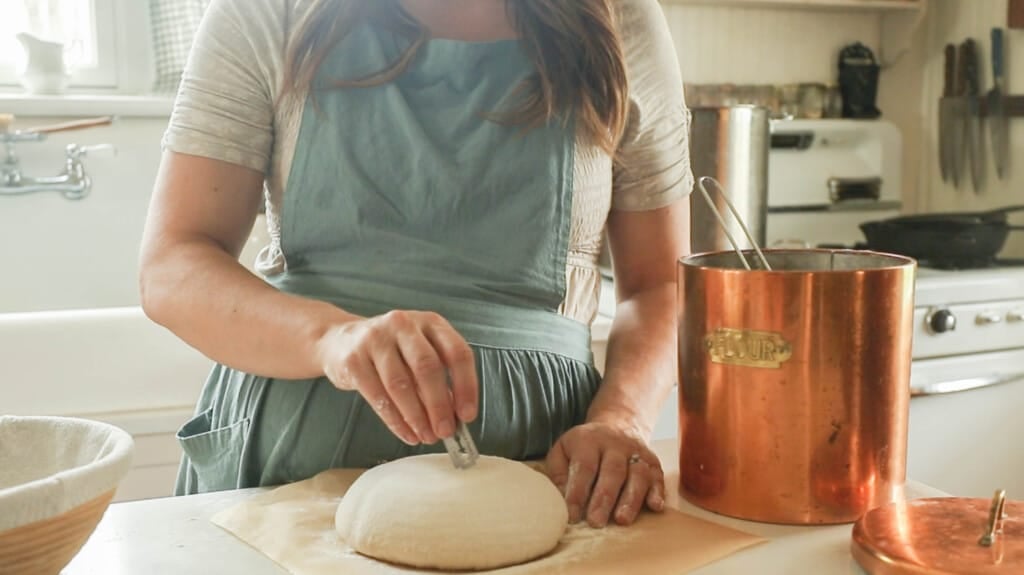 a woman wearing a green apron scoring a sourdough boule on a countertop with parchment paper