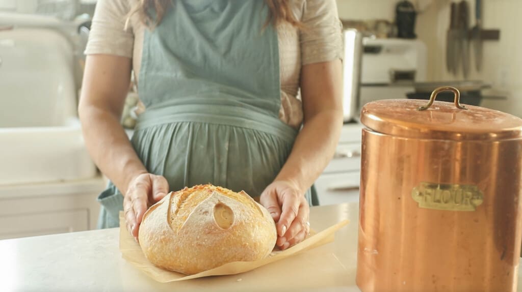 a woman wearing a green apron holding a loaf of sourdough boule on parchment paper on a white countertop