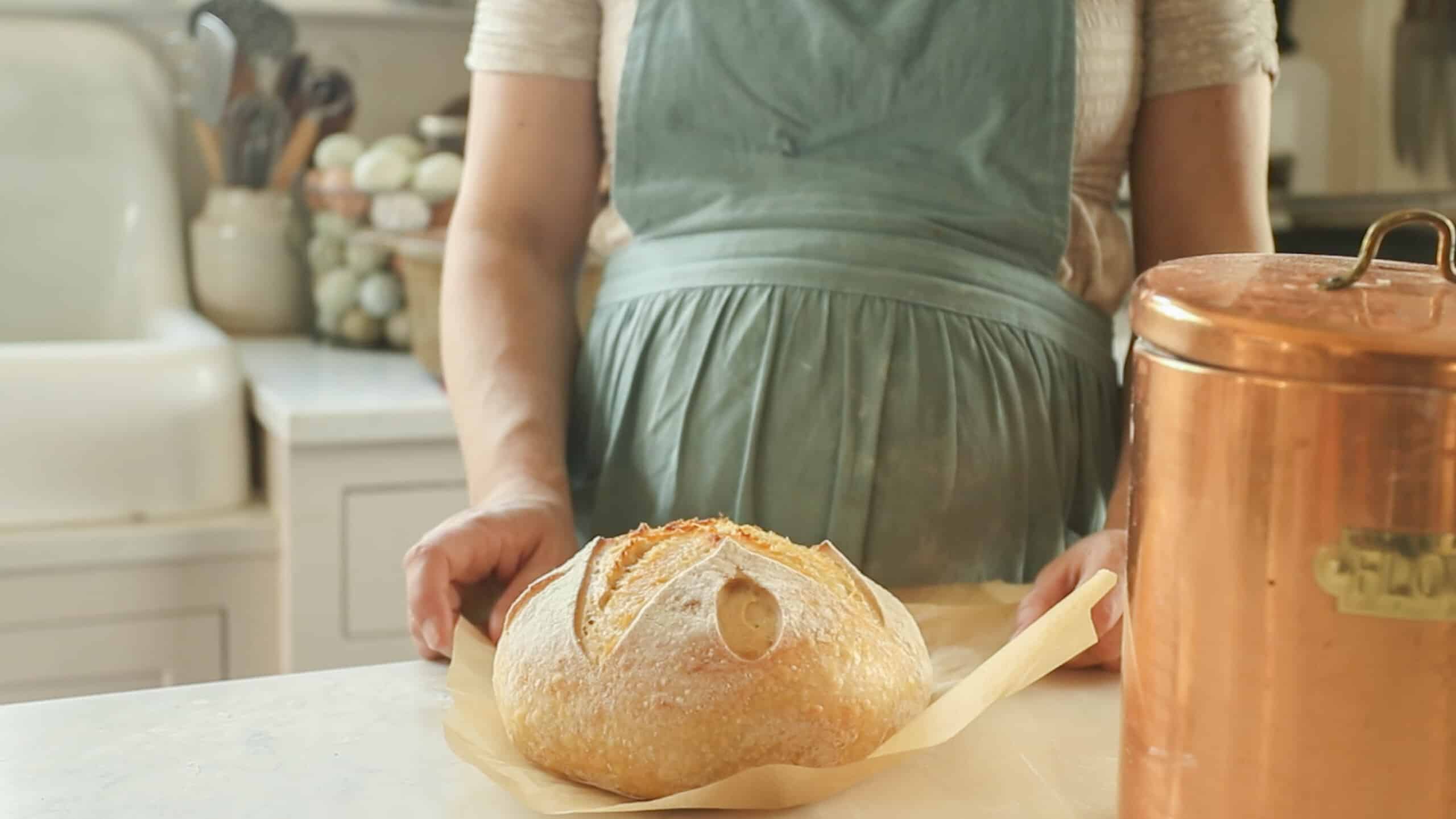 a sourdough boule on parchment paper on a white countertop with a woman in a green apron. With a copper canister to the left