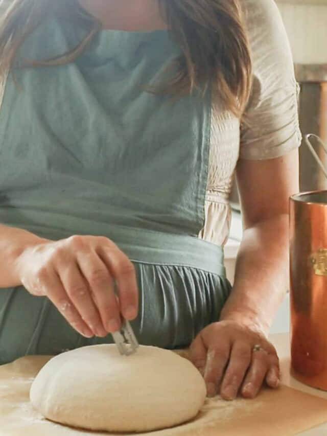 a woman wearing a green apron scoring a sourdough boule on a countertop with parchment paper