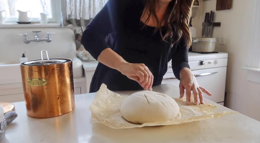 woman scoring sourdough boule dough on parchment paper on a white counter