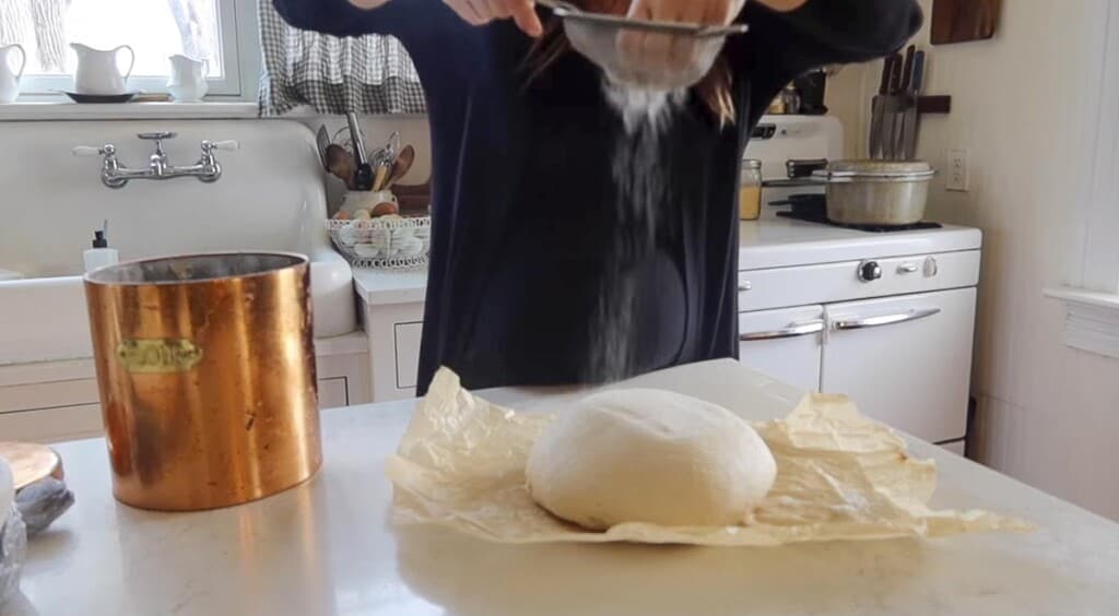a woman dusting flour onto a loaf of bread dough on parchment paper
