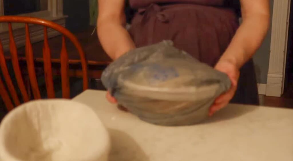 a banneton basket full of dough in a plastic grocery bag on a white countertop