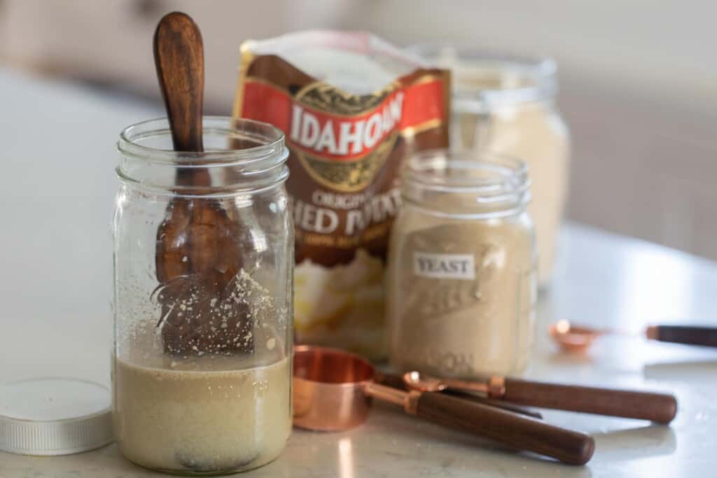 potato flake sourdough starter ingredients next to a jar of mixed starter with a wooden spoon inside