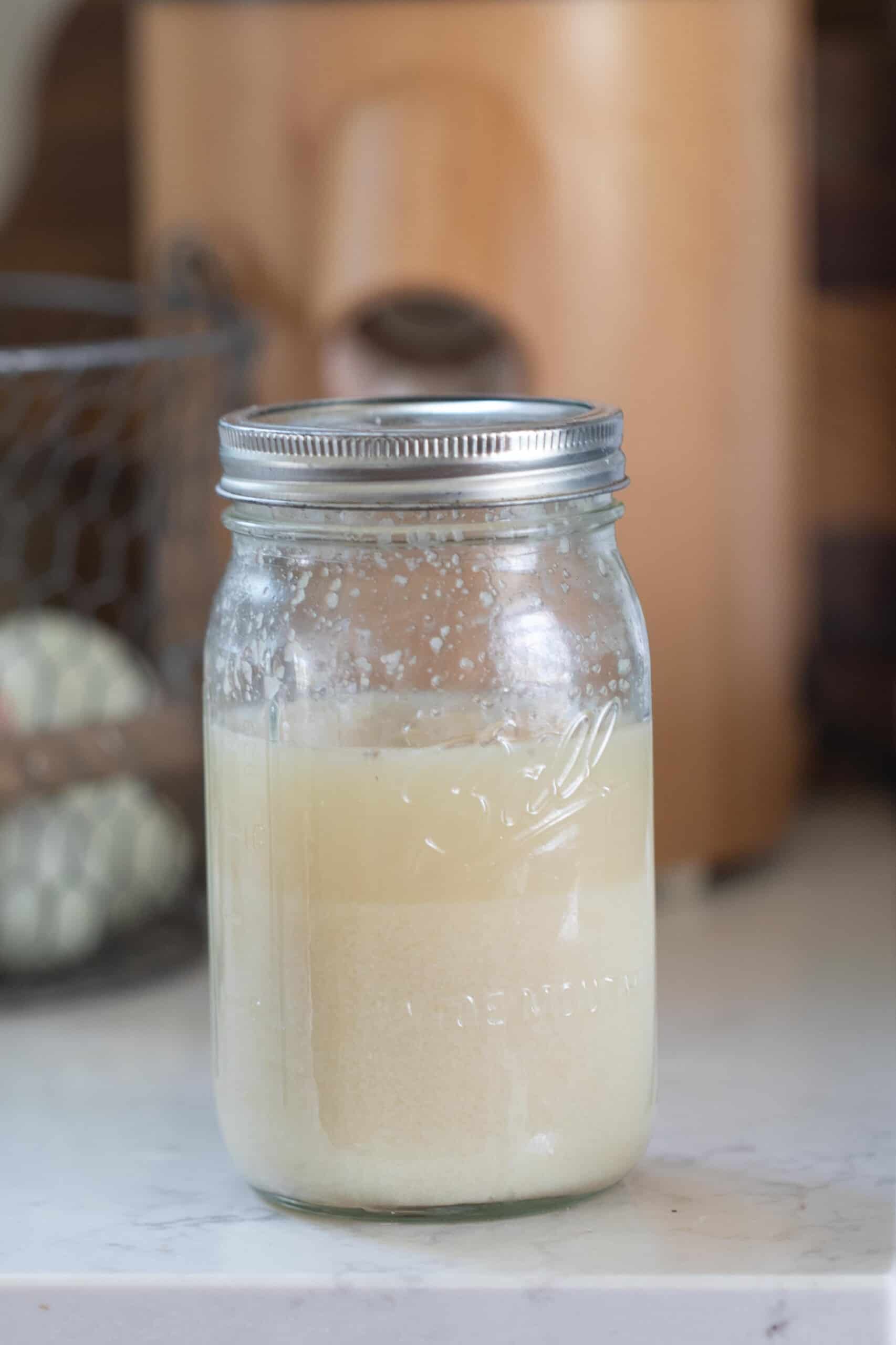 A glass jar of potato flake sourdough starter with a grain mill and eggs in the background on a white counter top