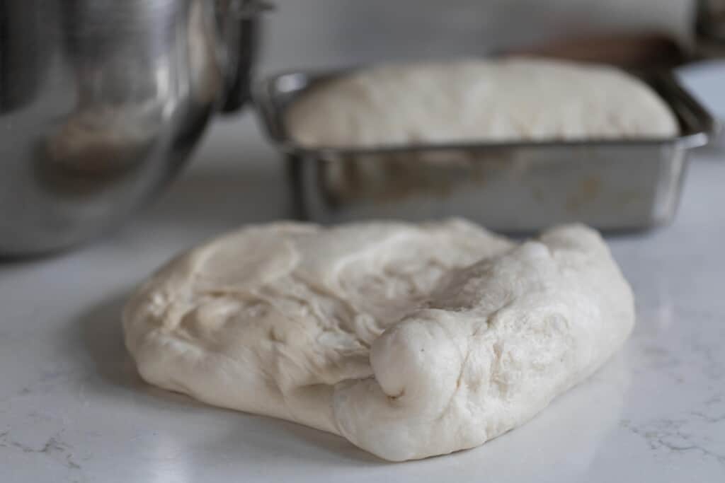 One loaf pan of potato flake sourdough starter sandwich bread with another dough ball being shaped and prepared for a loaf pan