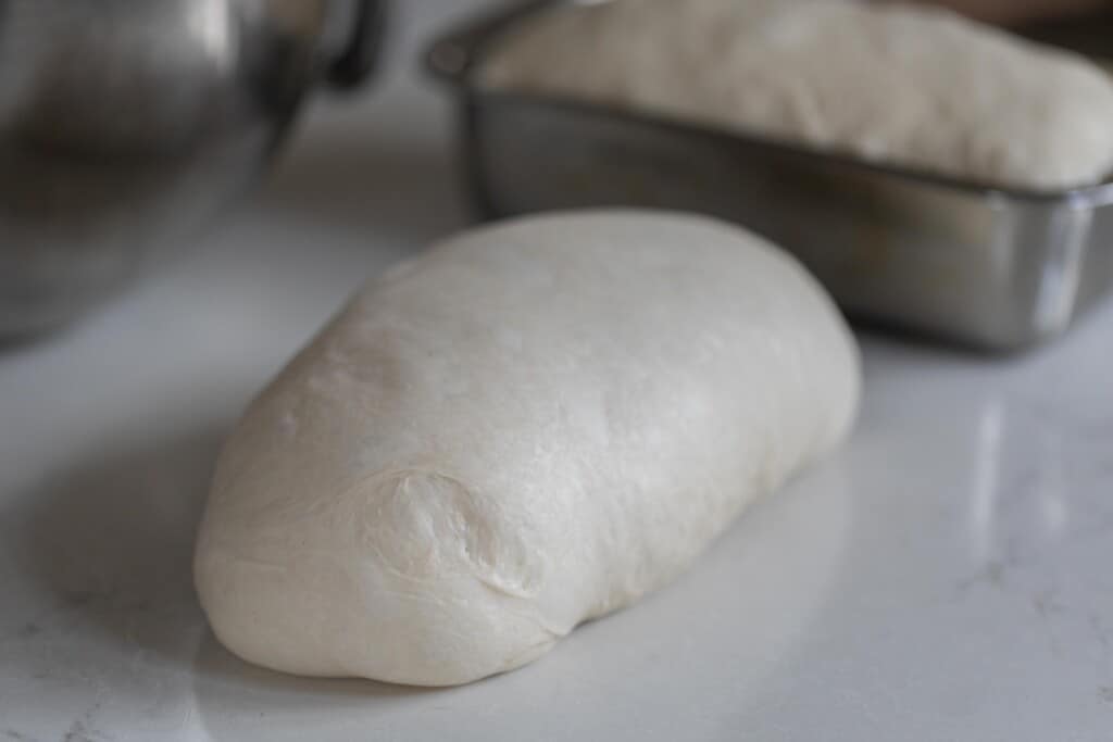 Potato flake sourdough starter sandwich bread rolled up and shaped on a white counter top with another loaf in the tin loaf pan in the background