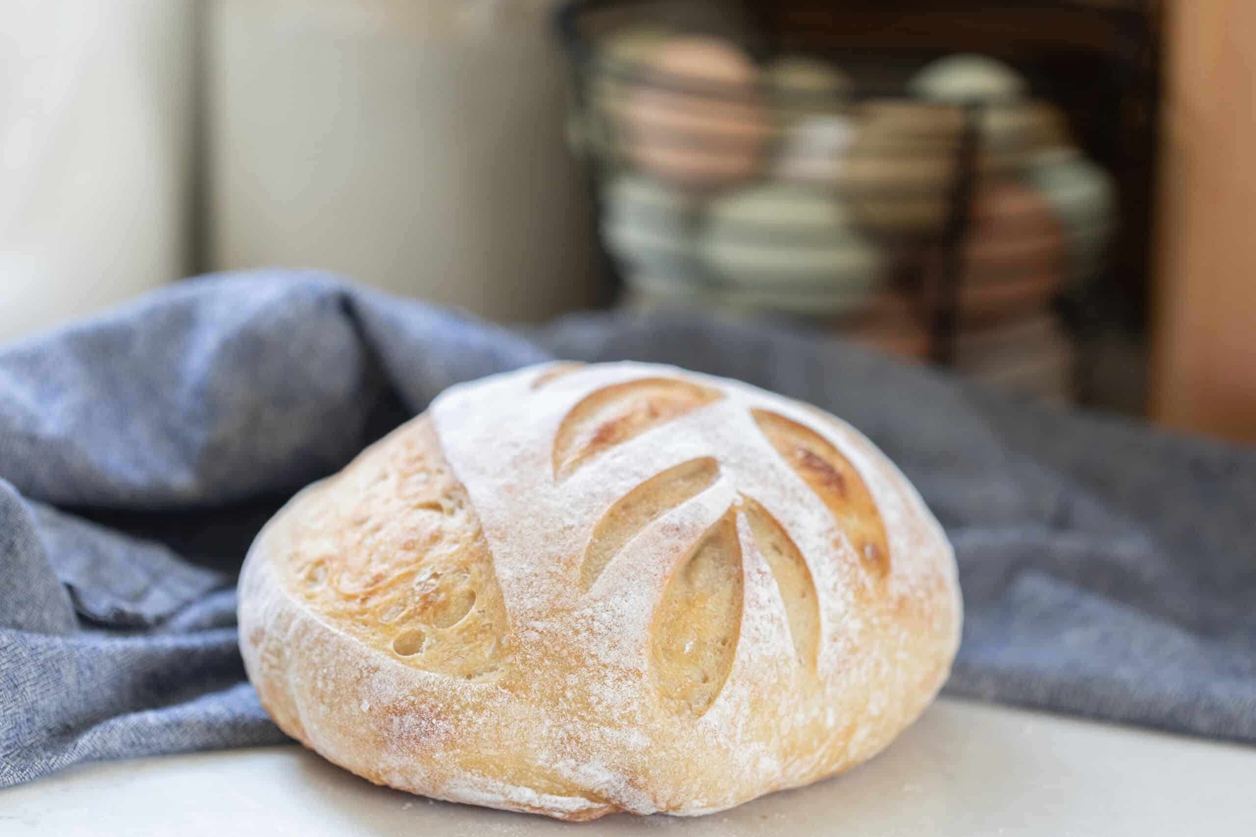 loaf of same day sourdough bread with a leaf pattern on a white counter with a blue towel and a basket of eggs in the background