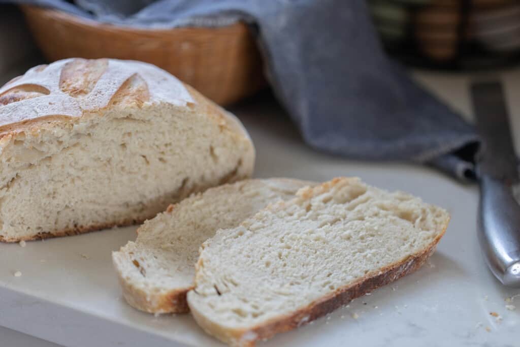 loaf of same day sourdough boule with two slices laying to the right on a white countertop. A blue towel in a basket is behind the loaf