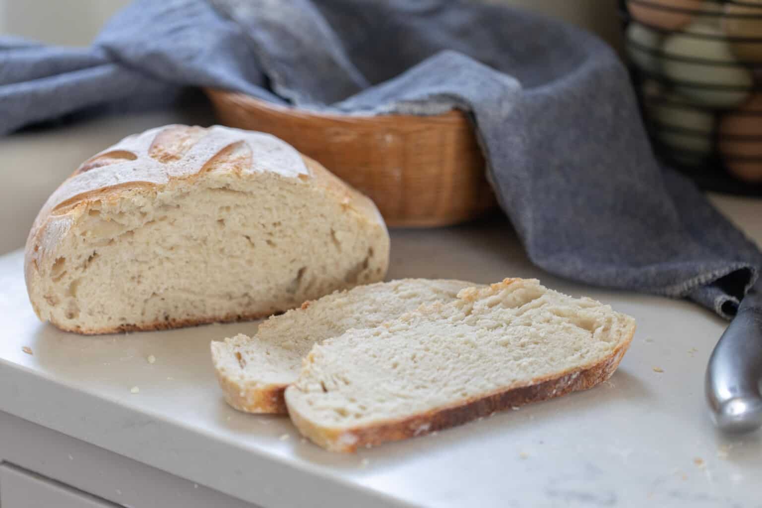 a sourdough boule with two slices sliced off and laying next to the loaf on a white countertop. A basket with a blue towel is in the background