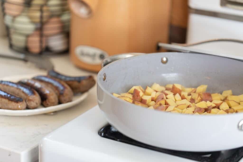 Cubed potatoes cooking in a gray large skillet on a white gas stove top with cooked sausage on a platter on the side