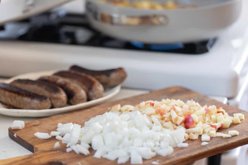 Cooked smoked sausages on a platter with a cutting board in front with diced onion and apples