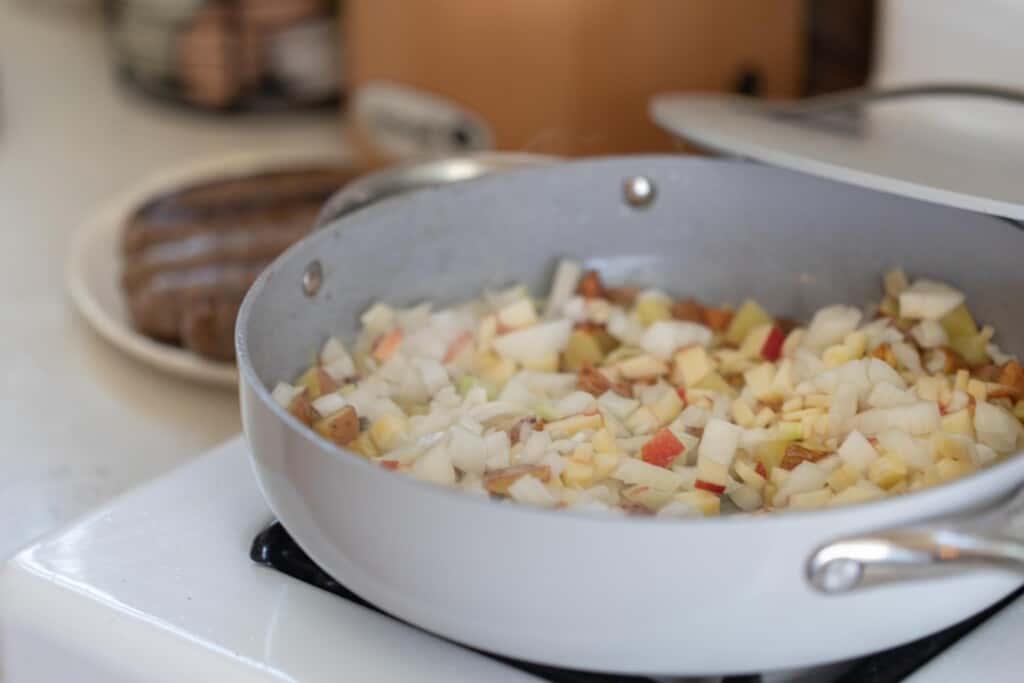 A large gray skillet on a white gas stove top with potatoes, onions, and apples 