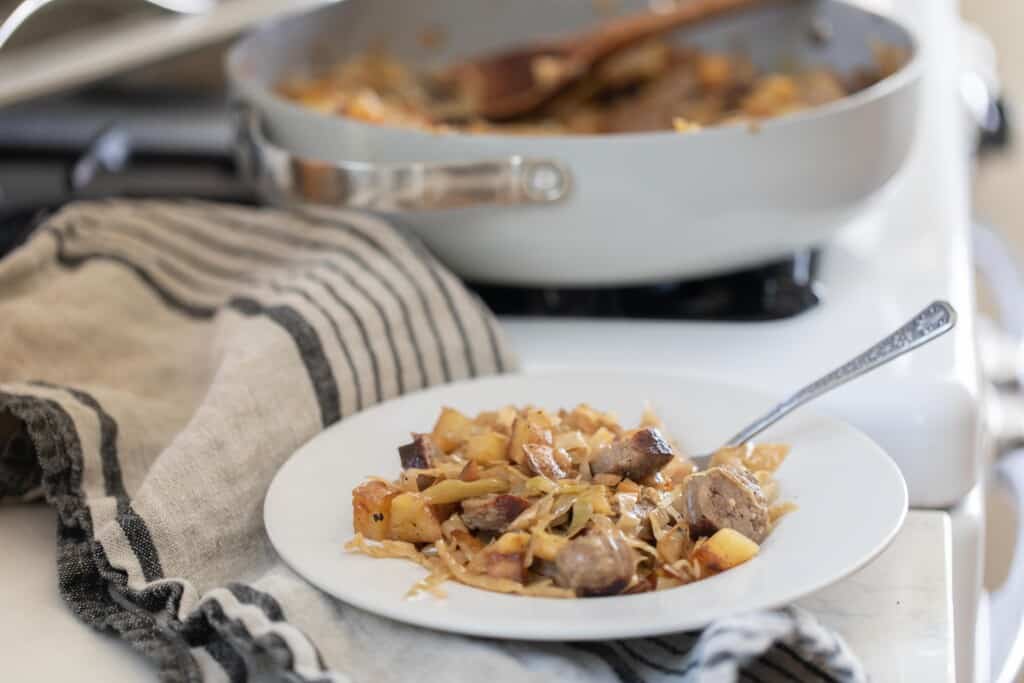 A bowl of sausage and sauerkraut with a fork and a skillet with the rest of the dish inside