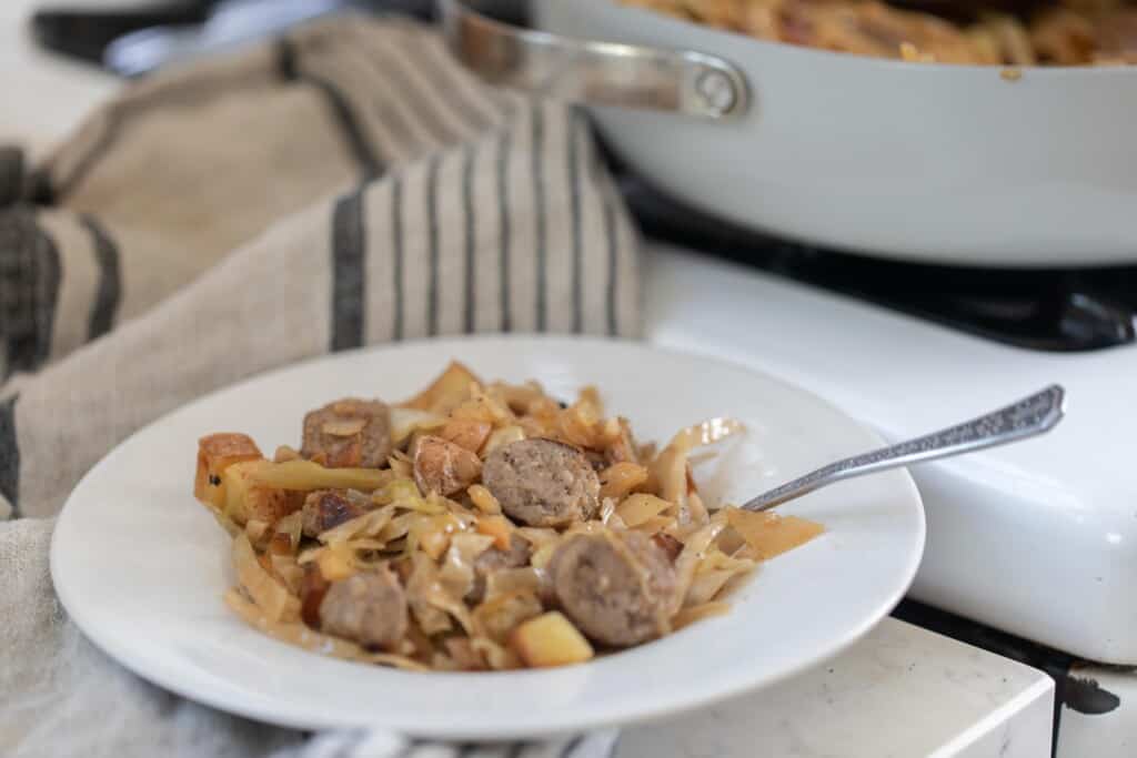 A white bowl filled with sausage and sauerkraut with a silver fork in it and a gray skillet in the background and a white and blue tea towel