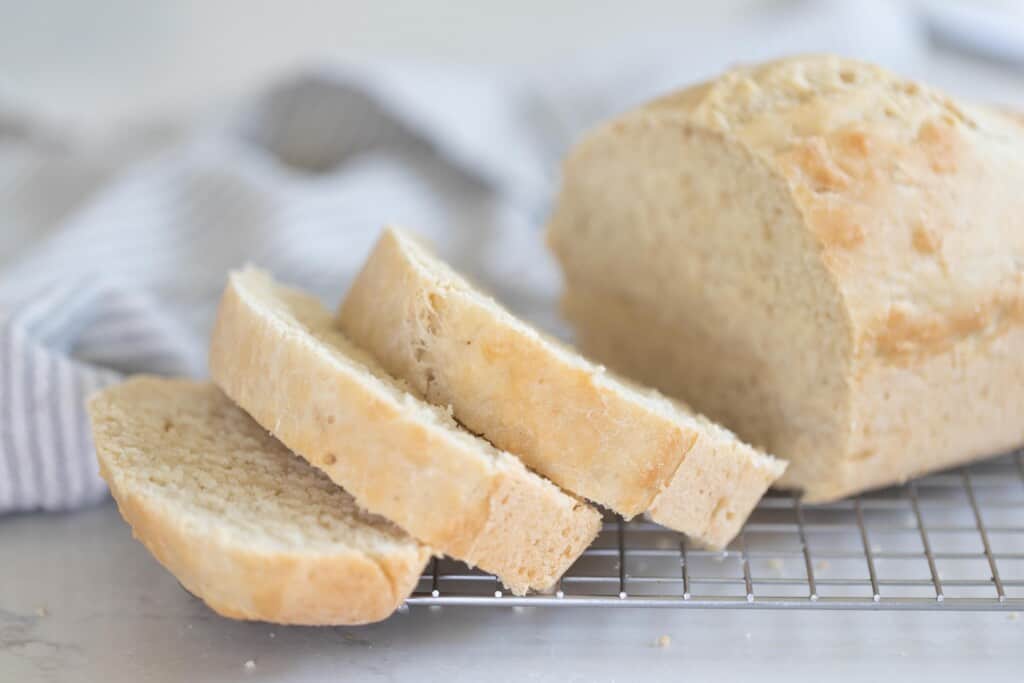 a loaf of sourdough beer bread with three slices cut, sitting on a wire rack with a blue and white tea towel in the background