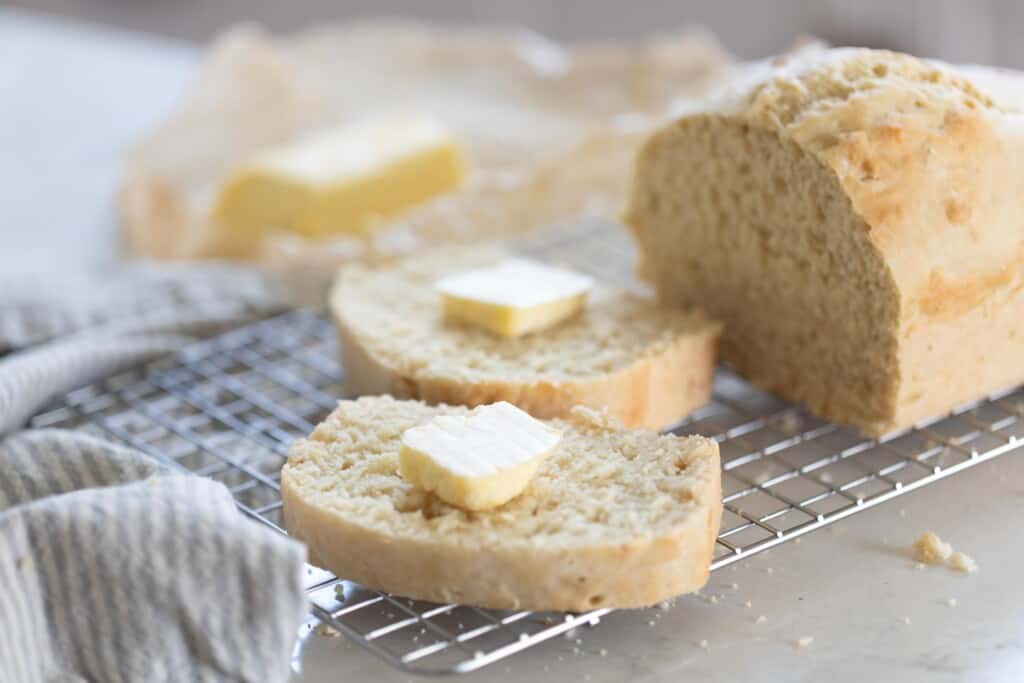 a loaf of sourdough beer bread on a wire rack with two slices of bread with butter next to the loaf.