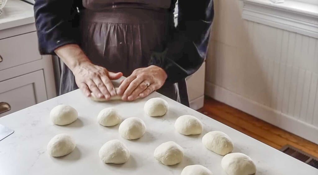 a woman wearing a gray apron rolling out dough on a white countertop with dough balls all around the counter