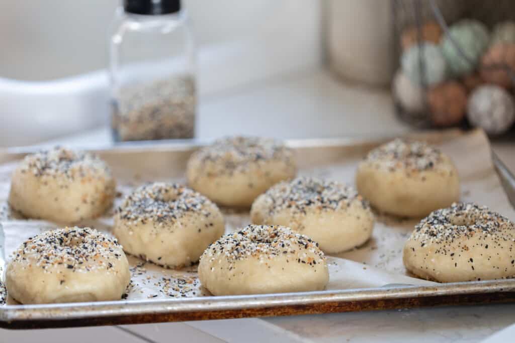 sourdough bagels topped with everything seasoning on a parchment line baking sheet ready for the oven