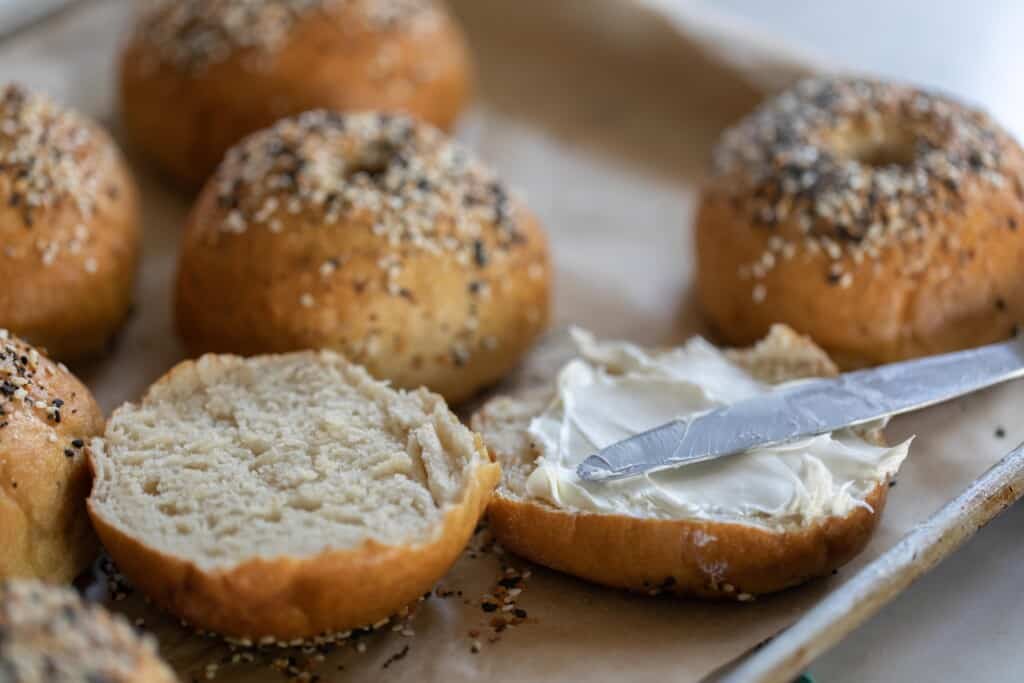 a sourdough bagels sliced in half and smothered with cream cheese and a knife resting on the bagel. More bagels in the background