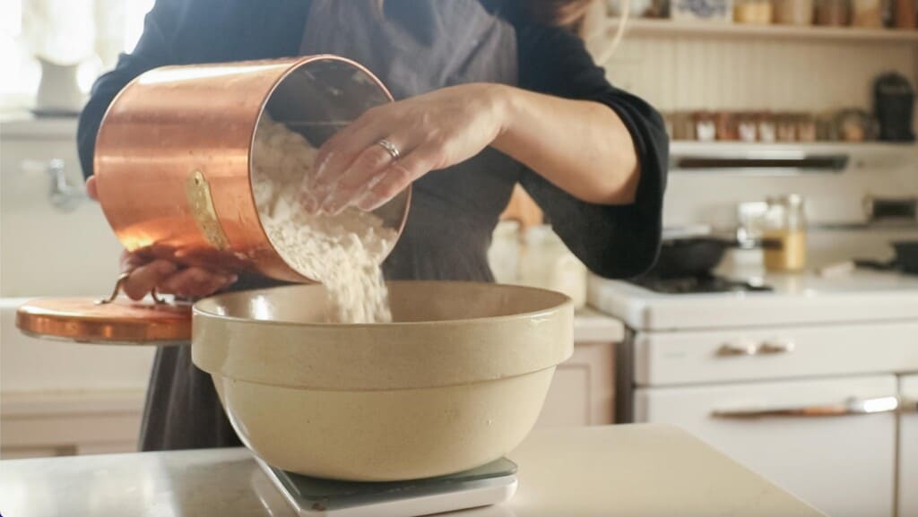 woman pouring flour into a bowl over a scale in a white kitchen