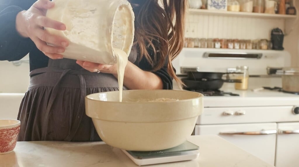 a women wearing a gray apron adding sourdough starter to a bowl on a white countertop