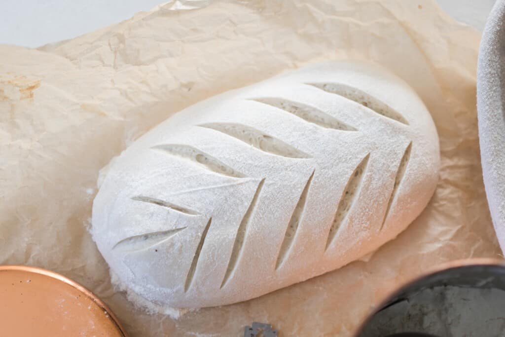 overhead photo of a rectangular shaped loaf of sourdough bread with a beautiful wheat pattern scored on top. The dough sits on parchment paper