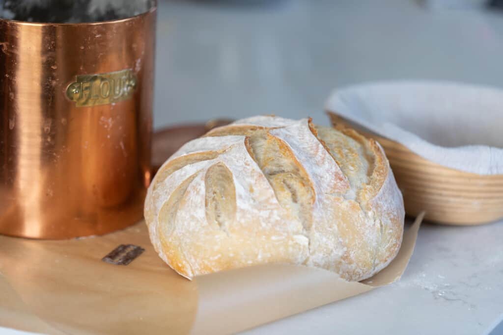 a homemade boule of easy sourdough bread on parchment paper on a white counter. A copped container and banneton basket is behind the loaf